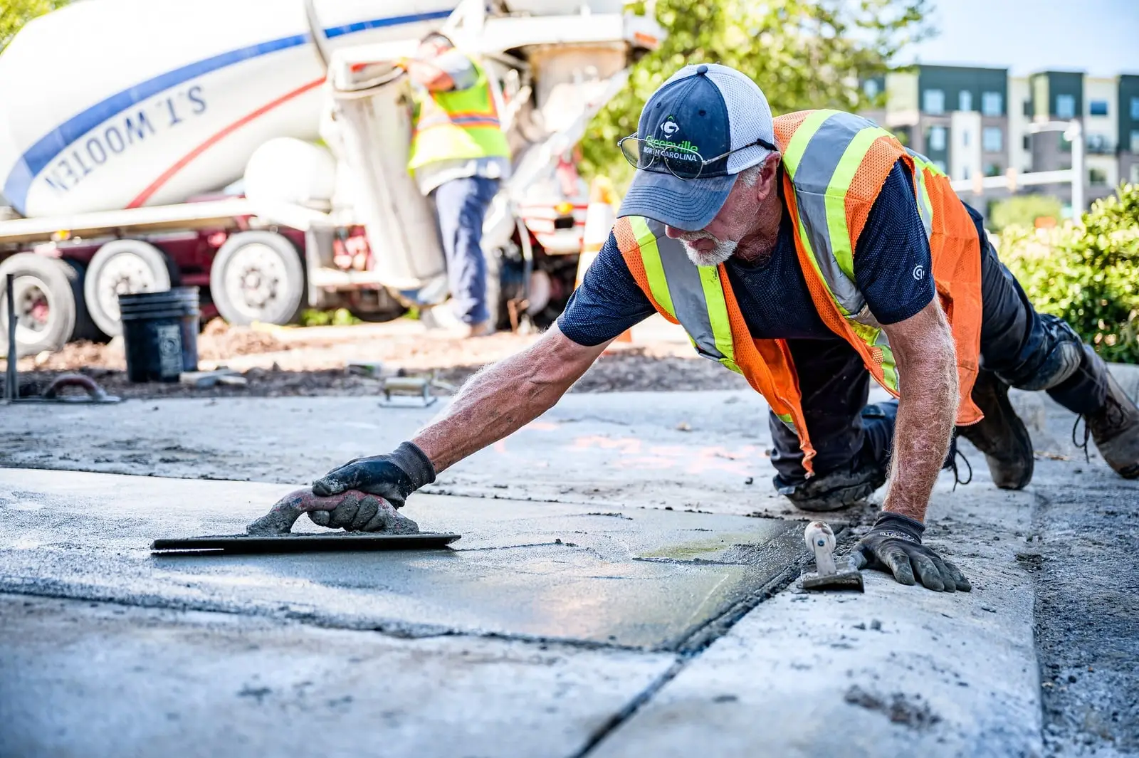 Concrete finisher hand-troweling the edge of a new sidewalk section next to a concrete mixer truck