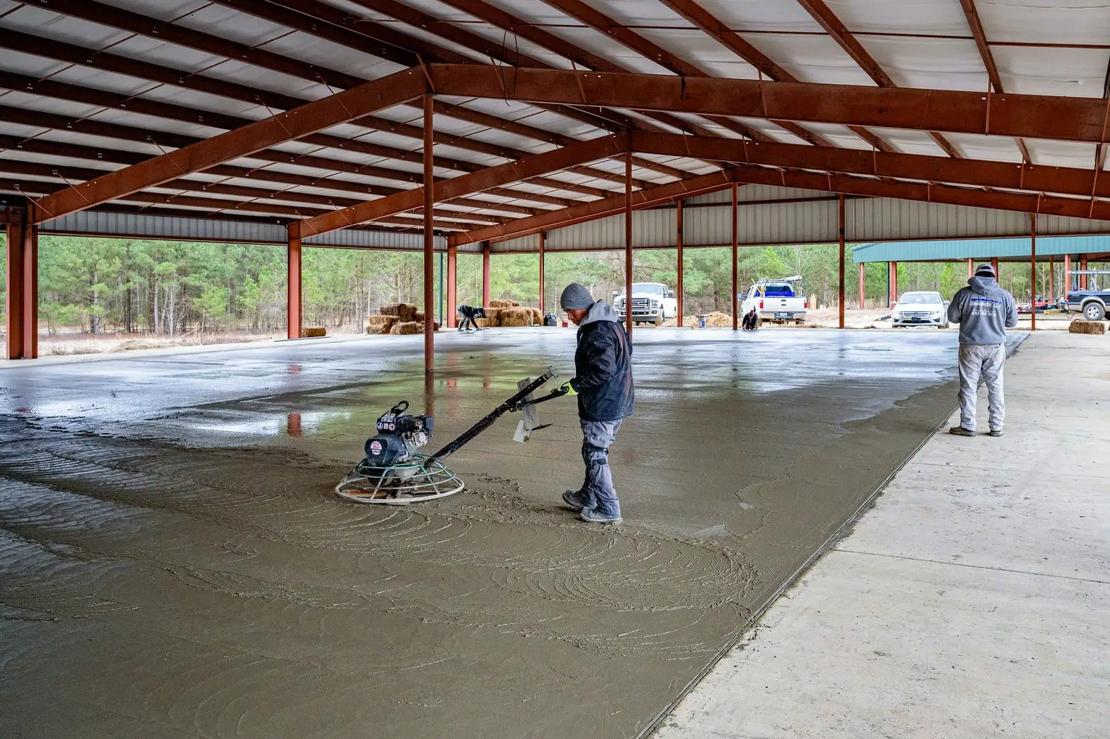 Worker using a power trowel to finish a large concrete slab under a steel pavilion roof